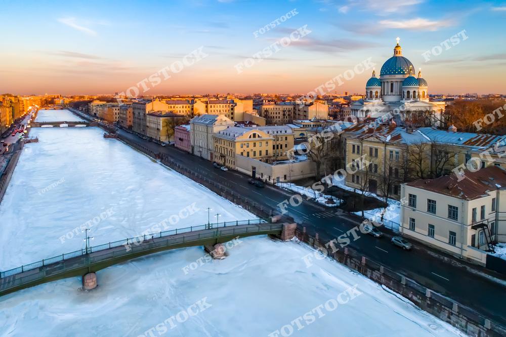 На фотографии изображен Красноармейский мост, пересекающий реку Фонтанку, покрытую льдом в зимний период. На заднем плане виден Троицкий собор, выделяющийся своими голубыми куполами с золотыми звездами. Утренний свет подчеркивает архитектурную гармонию исторических зданий и современный облик моста. Этот вид демонстрирует красоту и величие зимнего Петербурга.